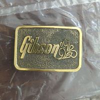 Top view of Gibson belt buckle showing gold-tone Gibson script and decorative flourish on textured black background.