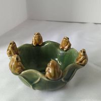 Close-up photo showing green ceramic bowl with seven brownish-green frog figurines evenly spaced on the rim, glossy finish.