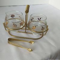 Front view of MCM cream and sugar set in gold tone wire holder with matching tongs on white surface.