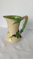 Front and side view of the ceramic pouring jug with colorful bird handle and flower spout, showing cream base, green interior, and condition features.
