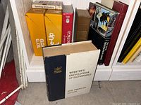 Books on a white shelf including Webster's Encyclopedias of Dictionaries with blue/white cover, and other assorted reference and atlas books with colorful spines including yellow and red covers