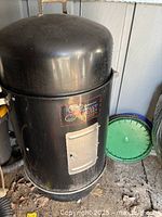 Full view of the large black cylindrical Brinkmann Gourmet Charcoal Smoker with dome lid, sitting outdoors on a dirt and stone surface.