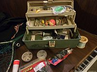 Open view of the green and beige Plano tackle box showing compartments filled with assorted fishing lures, bobbers, and accessories. Two fishing line spools and a package of red rubber pants are placed in front.