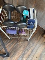 Metal two-tier rack with multiple dumbbells and a yoga block placed on a wooden floor against unfinished wall.