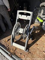 Metal hose holder reel positioned on ground with leaf debris around, showing two large circular spools and handle.