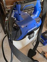 Side view of Kobalt 24V Max cordless garden sprayer showing blue and white tank, handle, and attached hose with spray nozzle holder strap.