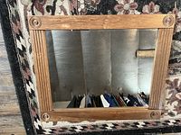 Square mirror with a light wooden frame featuring decorative carved rosettes at each corner, hung on a wall reflecting clothes.