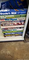 Photo showing a stacked collection of shooters catalogs and gun guides on a shelf including Shooter's Bible, Gun Trader's Guide, Gun Digest, Master Catalog, and Cartridges of the World.