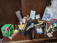Pile of kitchen utensils and gadgets on a dark wood surface, showing tape, ponchos in packages, various spatulas, tongs, and corn cob holders.