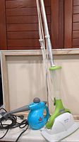 Blue Bissell Steam Shot handheld steamer and green and white Smart Living steam mop side by side on a counter.