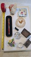Overview photo of all items including Coca-Cola pen, porcelain chicken, floral plate, trivet, figurines, glass inkwell/candle holder, picture frame, and ceramic flowers.