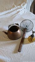 Side view of three items: copper kettle, vintage metal mesh strainer with wooden handle, and yellow glass soap bottle with metal pump.