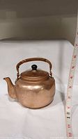 Copper teapot with woven handle and black knob on lid, front side view with measuring tape showing size