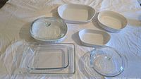 Set of 7 cooking casserole dishes laid out on white fabric, includes various ceramic and glass dishes with lids and without, all appear lightly used