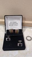 Two pairs of cufflinks and one spinning ring inside a black jewelry box with white interior marked 'Di Capri Collection Milan Italy'.