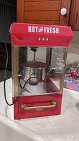 Front angle showing the popcorn maker with red top and 'Hot and Fresh' branding, glass housing, and power cord.