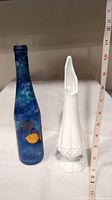 Two glass items shown side by side: a white milk glass vase with a hobnail pattern and a tall blue bottle decorated with painted fish and underwater themes. A measuring tape is visible for scale, showing heights around 10-12 inches.