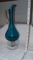 Tall blue teardrop-shaped glass vase with clear handle and pedestal base, displayed next to a measuring tape showing approximately 11 inches tall.