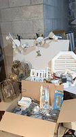 Overview of lot showing two white multi-bulb light fixtures, various clear lantern-style light covers, white plastic light switch plates, a white slatted wood item, and boxes containing assorted plumbing and hardware parts.