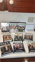 View of nine DVD box sets for M*A*S*H TV series arranged on a kitchen counter showing cover artwork and titles.