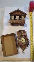 Photo showing both the mini cuckoo clock and Swiss carved wind-up clock next to a tape measure for size reference, with the cuckoo clock featuring figurines in the window and the wind-up clock having two pull chains.