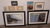 Seven framed pictures leaning against a wall showing two large Niagara Falls prints and five smaller miscellaneous framed pictures and documents.