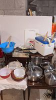 Photo showing a table with Pyrex baking dishes, steel pots with lids, a Coleman cooler, a Starbucks mug, wooden utensils, blue mixing bowl, and miscellaneous kitchen items.