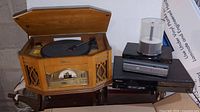 Photo of vintage wooden record player and radio combo with speakers, paired with a stack of modern electronic devices including a CD player and DVD player, plus spindle of blank CDs.