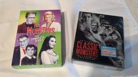 The Munsters Complete Series DVD box set next to The Classic Monsters Collection Blu-ray box set on a white surface.