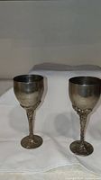 Two silver plated goblets standing side by side displaying grape vine themed stem and base details