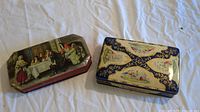 Two vintage tin cans on a white cloth, one with William Tell scene and one with floral and bird motifs