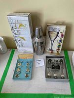 Full view of all 5 bar accessory items arranged on a flat surface including cocktail shaker, shot glasses, glass charms, wine markers and stirrers