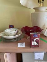 Display of vintage ruby red glass pedestal bowl with assorted dishes and boxed napkin rings on wooden shelf.
