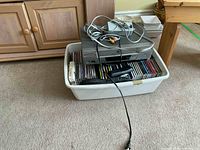Photo showing Philips DVD player and VCR player stacked on top of a white plastic bin filled with music CDs and audio cables on top. Carpeted floor and wooden furniture in background.