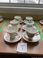 Photo showing six fine china teacups with floral patterns and matching saucers arranged side by side on a windowsill, each cup has a unique shape with gold rims and handles.