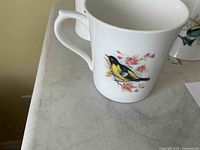 Side view of a white tea or coffee mug showing a colorful bird and floral motif typical of the Queens Birds of America series.
