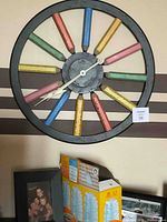 Round metal framed wall clock with colored pencil design for hour markers, mounted on a wall above a small shelving unit.