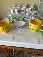 Photo showing large yellow smiley face mugs, white cups with floral and bird prints, saucers, and a green floral teacup set on table.