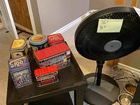 Photo showing six collectible Cadbury tins with vintage designs and two red double-decker bus shaped tins on a small dark table, next to a black Comfortmate pedestal fan with a note indicating 35" by 16" measurements.