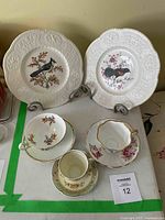 Two large white porcelain collector plates each with a bird design in the center and scalloped lacy edges displayed on metal plate stands along with three floral decorated porcelain teacups and matching saucers arranged on a white surface.