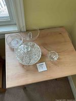 Six crystal and glass items on wooden table including a large round clear glass plate, crystal candy dish with decorative patterns, smaller crystal bowl, additional glass bowls, and two hexagonal candle holder bowls.