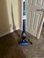 Full view of blue Electrolux Ergorapido cordless stick vacuum cleaner upright on carpet, showing main body, handle, power head, and power cord.