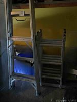 Two aluminum ladders standing in a garage or storage area: one 12.5-foot folding multi-purpose ladder with visible locking joints and platform, and one 6-foot standard step ladder.