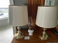 Two brass table lamps with white fabric lampshades and a white ceramic oil lantern between them on a wooden surface.