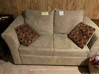 Front view of beige fabric loveseat with two large beige back cushions and two smaller patterned accent cushions, placed in a wood-paneled room.