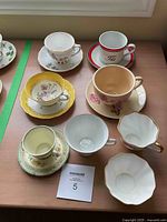 Top view of 7 assorted fine china teacups and their matching saucers arranged on a wooden table, including a yellow cup, floral patterns, and a red banded cup.