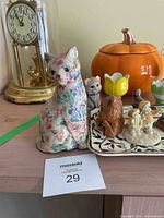 Front view of ceramic cat, ceramic pumpkin container, smaller cat figurine, flower-shaped egg cups, figurines on decorative tray, on wooden table with clock in background.