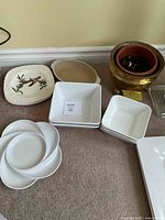 Shows various ceramic dishes of different shapes and sizes arranged on floor including square bowls, decorative plate, oval dish, round plates, and copper pot.