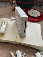 White Nintendo Wii console vertically positioned on grey stand beside white Wii Balance Board on carpet, with some household items in background.