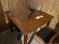 Solid wood square table showing wear and scratches on surface, positioned against wood-paneled wall.